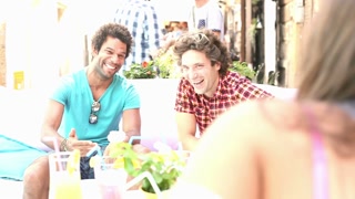 Two young, male friends sitting and chatting with friends on terrace cafe in town in summer, graded warmer.